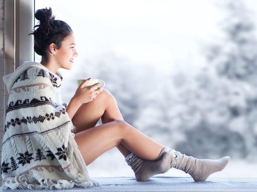 woman drinking coffee wearing knitted nordic print poncho sitting home by the window